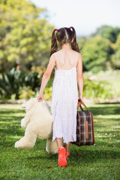 Niña caminando en el parque con una maleta y un oso de peluche