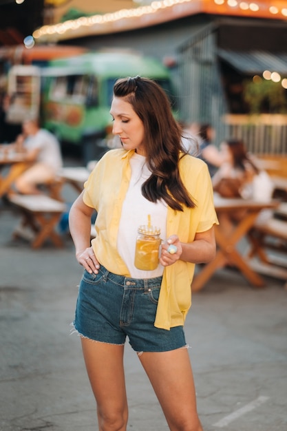 Una niña con una camisa amarilla y pantalones cortos con un vaso de jugo de limón está parada en la calle.