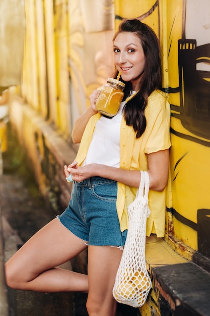 Una niña con una camisa amarilla, pantalones cortos y zapatos negros con una red de limones y jugo de limón camina por una calle de la ciudad. Una mujer con un cóctel en sus manos.