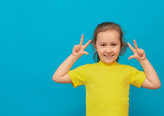 Una niña con una camiseta amarilla mostrando tres dedos en sus manos sobre fondo azul.