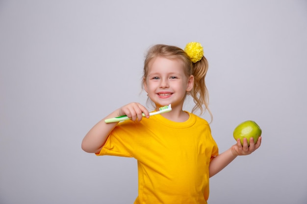 Una niña con una camiseta amarilla sostiene un cepillo de dientes y una manzana en un fondo blanco