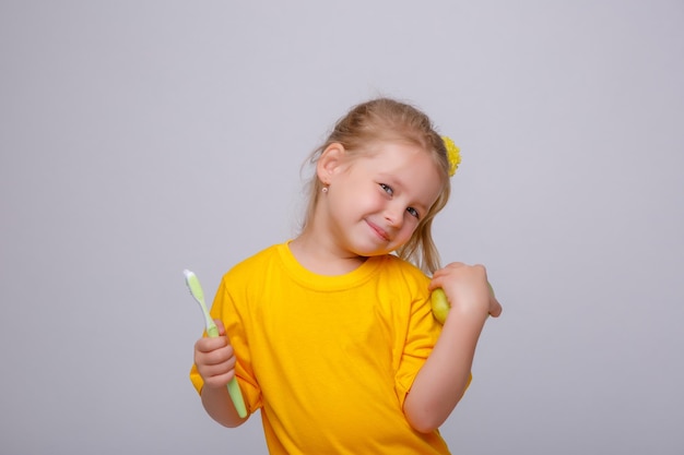 Una niña con una camiseta amarilla sostiene un cepillo de dientes y una manzana en un fondo blanco