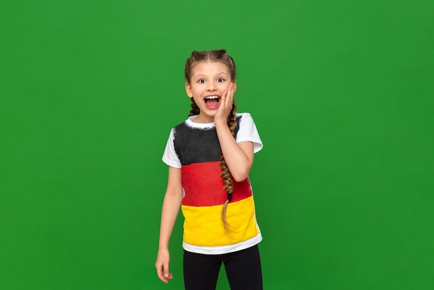 Una niña en una camiseta con una bandera alemana sonríe sorprendida sobre un fondo verde aislado Cursos de alemán para escolares