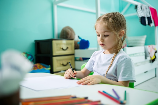 Una niña con una camiseta blanca se sienta en su habitación a la mesa y dibuja con lápices de colores