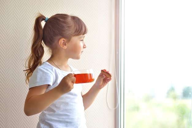 Una niña con una camiseta blanca sostiene una taza de té y un hígado en forma de corazón rojo de pie junto a la ventana, mirando. delicioso bocadillo dulce. seguro en casa, autoaislamiento durante una pandemia.