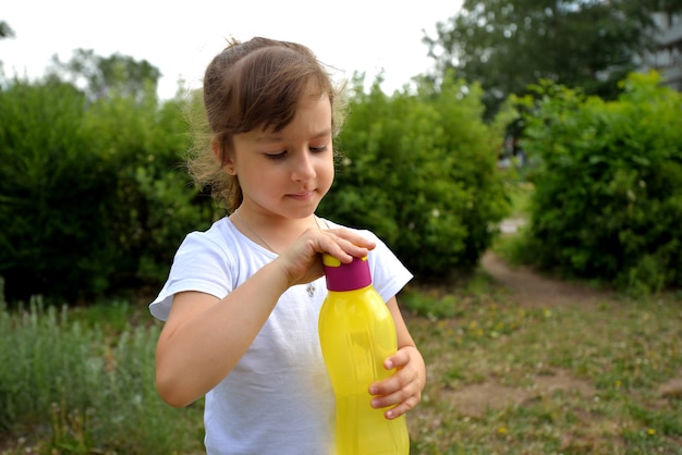 Una niña con una camiseta blanca en el verano en la calle bebe agua de una botella de plástico amarilla de calidad ecológica.