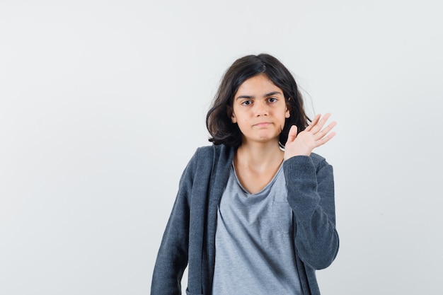 Niña en camiseta, chaqueta agitando la mano y mirando confiada, vista frontal.