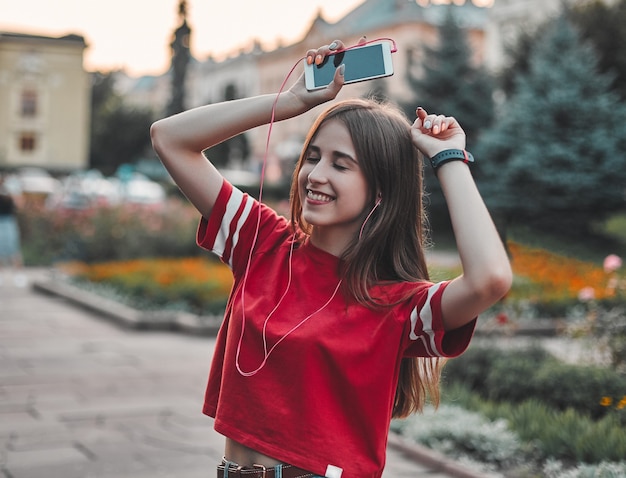 Una niña con una camiseta roja que usa audífonos con un teléfono en la mano está disfrutando de la música, cerrando los ojos y sonriendo