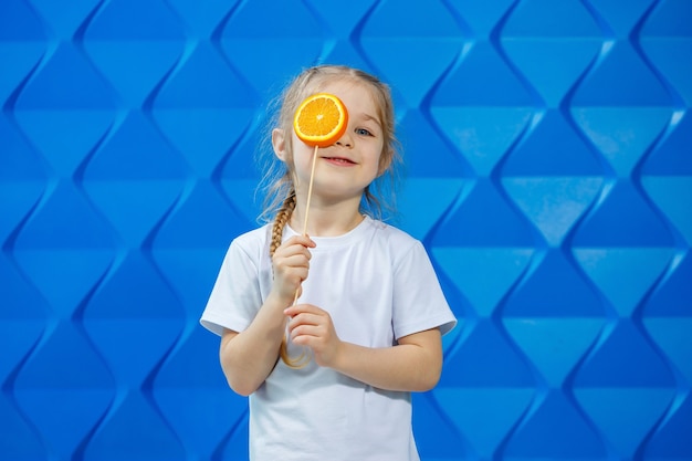 Una niña con una camiseta sonríe y sostiene dos mitades de fruta naranja frente a sus ojos sobre un fondo azul. Concepto: la lucha contra la avitomnosis primaveral.