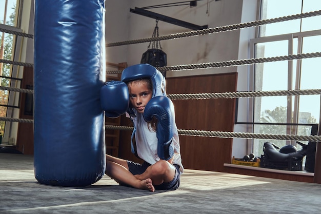 Una niña cansada con casco azul descansa en el ring al lado del saco de boxeo.