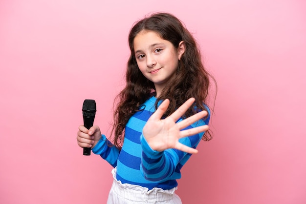 Niña cantante recogiendo un micrófono aislado en un fondo rosa saludando con la mano con expresión feliz