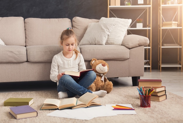 Niña en casa con libro y su juguete favorito, sentada en la alfombra. Niño leyendo historias emocionantes, sentado junto a un oso de peluche. Divirtiéndose y concepto de educación lúdica fácil, espacio de copia