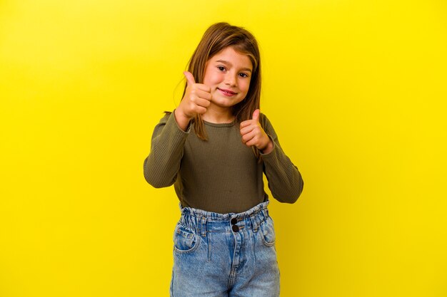 Niña caucásica aislada en amarillo levantando ambos pulgares, sonriente y confiada.