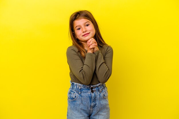 Niña caucásica aislada en amarillo mantiene las manos debajo de la barbilla, está mirando felizmente a un lado.