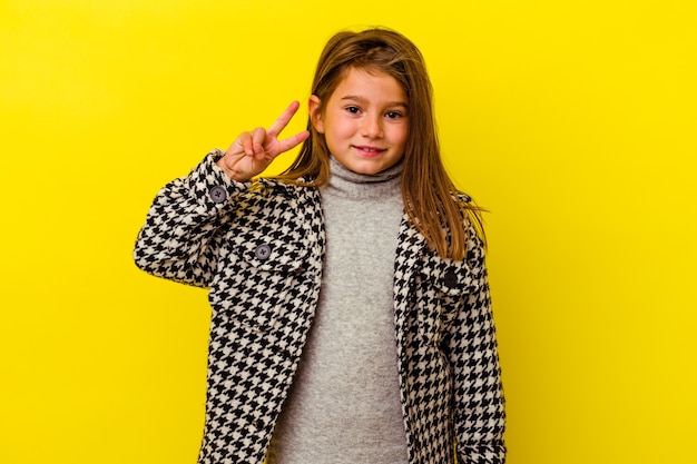 Niña caucásica aislada en amarillo mostrando el signo de la victoria y sonriendo ampliamente.