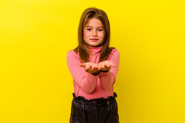 Niña caucásica aislada en amarillo sosteniendo algo con palmas, ofreciendo a la cámara.