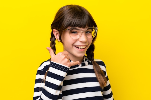 Niña caucásica aislada de fondo amarillo con gafas y haciendo gestos telefónicos