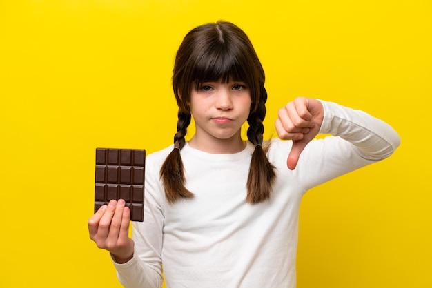 Niña caucásica aislada de fondo amarillo tomando una tableta de chocolate haciendo mala señal