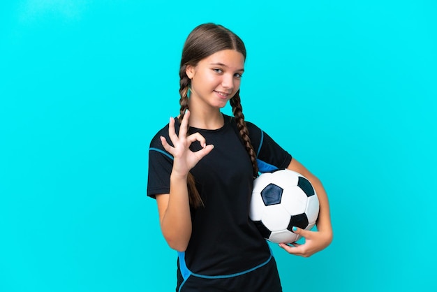 Niña caucásica aislada de fondo azul con pelota de fútbol y haciendo el signo de OK