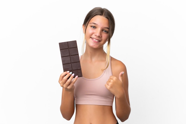 Niña caucásica aislada de fondo blanco tomando una tableta de chocolate y con el pulgar hacia arriba