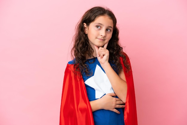 Niña caucásica aislada de fondo rosa con traje de superhéroe y pensando