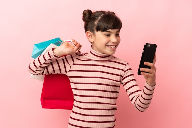 Niña caucásica aislada en la pared rosa sosteniendo bolsas de la compra y escribiendo un mensaje con su teléfono celular a un amigo