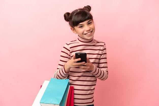 Niña caucásica aislada en rosa sosteniendo bolsas de la compra y escribiendo un mensaje con su teléfono celular a un amigo