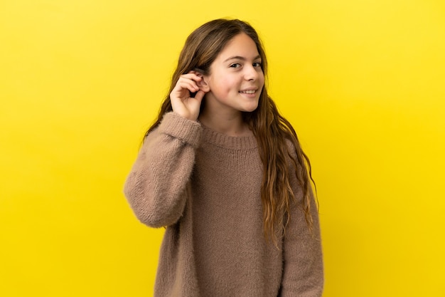 Niña caucásica aislada sobre fondo amarillo escuchando algo poniendo la mano en la oreja