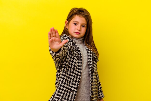 Niña caucásica aislada sobre fondo amarillo de pie con la mano extendida mostrando la señal de stop, impidiéndote.