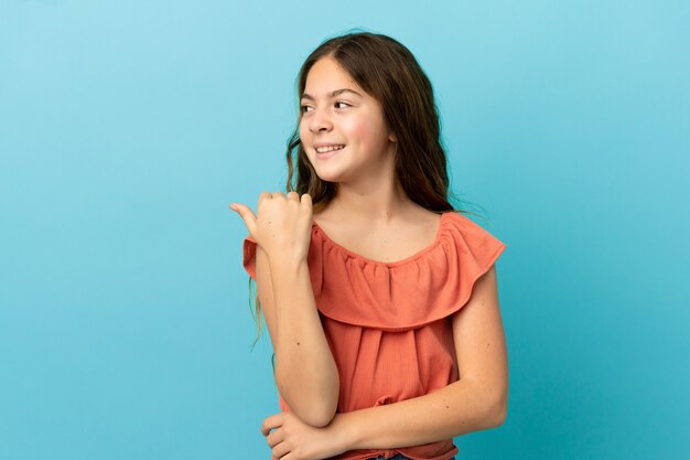 Niña caucásica aislada sobre fondo azul apuntando hacia el lado para presentar un producto