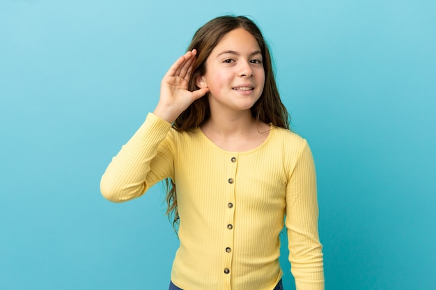 Niña caucásica aislada sobre fondo azul escuchando algo poniendo la mano en la oreja