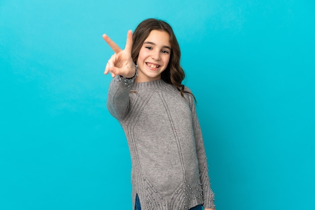 Niña caucásica aislada sobre fondo azul sonriendo y mostrando el signo de la victoria