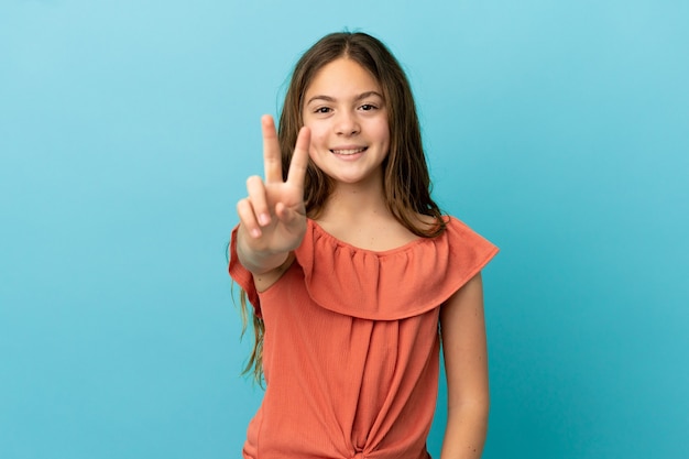 Niña caucásica aislada sobre fondo azul sonriendo y mostrando el signo de la victoria