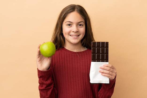 Niña caucásica aislada sobre fondo beige tomando una tableta de chocolate en una mano y una manzana en la otra