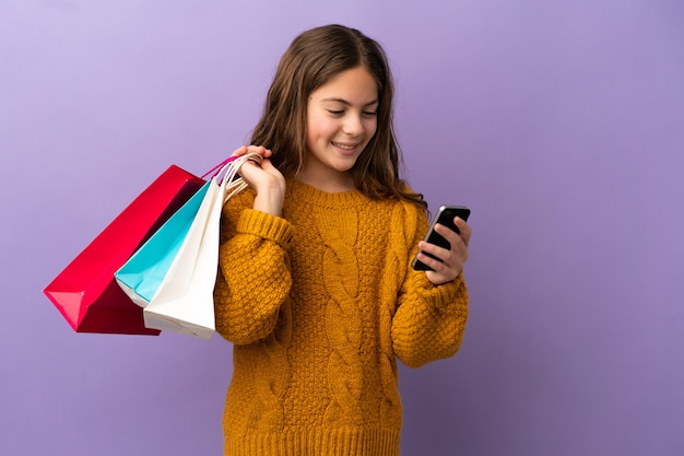 Niña caucásica aislada sobre fondo púrpura sosteniendo bolsas de la compra y escribiendo un mensaje con su teléfono celular a un amigo