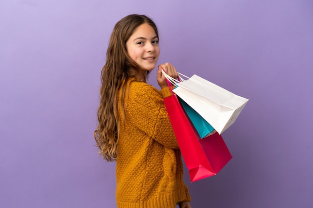 Niña caucásica aislada sobre fondo púrpura sosteniendo bolsas de compras y sonriendo