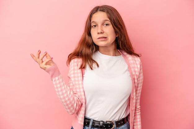 Niña caucásica aislada sobre fondo rosa dudando y encogiéndose de hombros en gesto de interrogación.