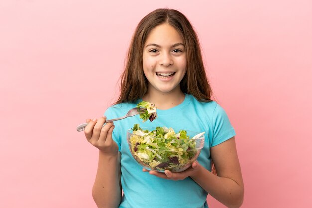 Niña caucásica aislada sobre fondo rosa sosteniendo un plato de ensalada con expresión feliz