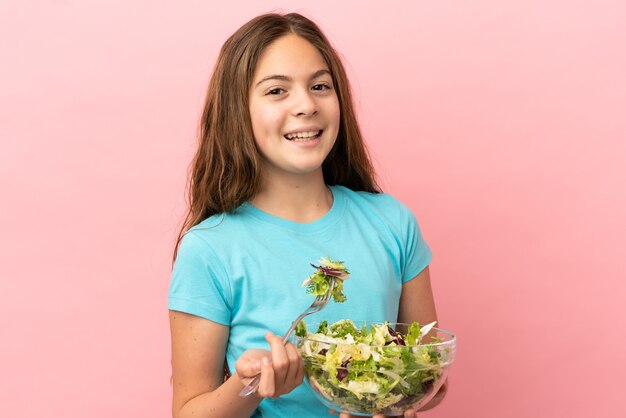 Niña caucásica aislada sobre fondo rosa sosteniendo un plato de ensalada con expresión feliz