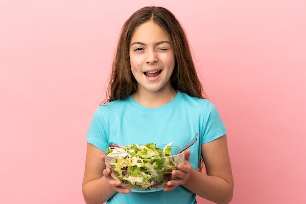 Niña caucásica aislada sobre fondo rosa sosteniendo un plato de ensalada mientras guiña un ojo