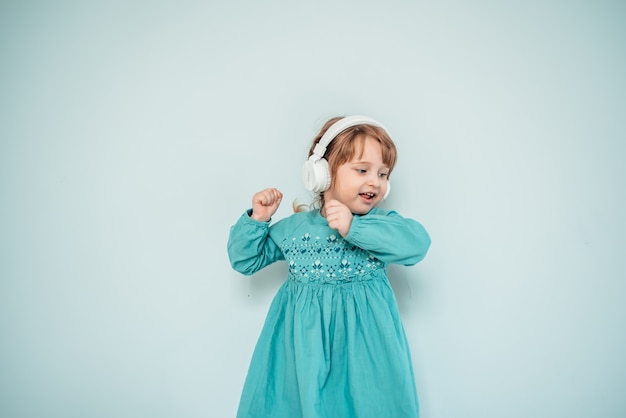 Niña caucásica en auriculares blancos escucha música y baila con un vestido azul turquesa brillante.