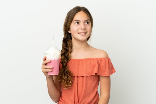 Niña caucásica con batido de fresa aislado sobre fondo blanco pensando en una idea mientras mira hacia arriba