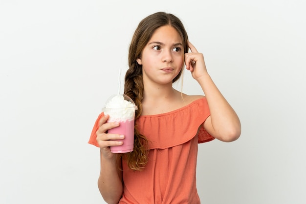 Niña caucásica con batido de fresa aislado sobre fondo blanco teniendo dudas y pensando