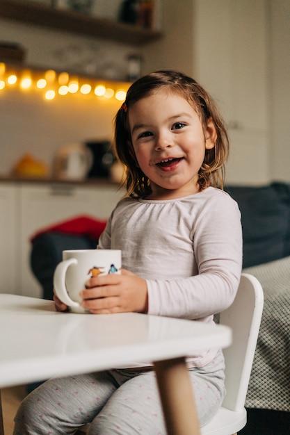 Niña caucásica bebiendo cacao de taza en casa. Luces de navidad bokeh