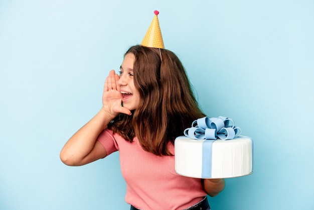 Niña caucásica celebrando su cumpleaños sosteniendo un pastel aislado sobre fondo azul gritando y sosteniendo la palma cerca de la boca abierta.