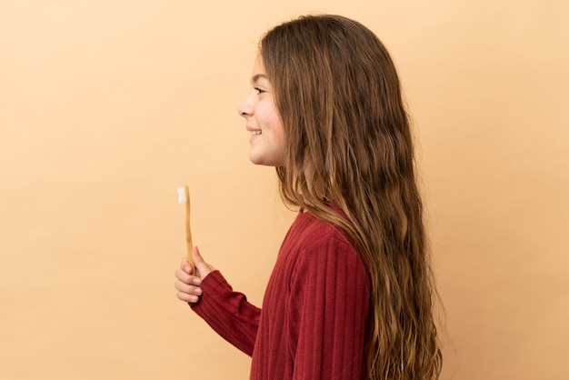 Foto niña caucásica cepillarse los dientes aislado sobre fondo beige riendo en posición lateral