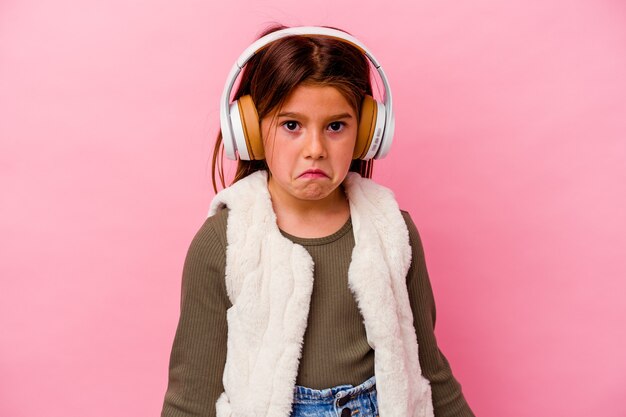 Niña caucásica escuchando música aislada en rosa encoge los hombros y abre los ojos confundidos.