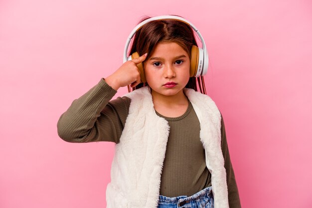 Niña caucásica escuchando música aislada en la sien que señala rosa con el dedo, pensando, centrado en una tarea.