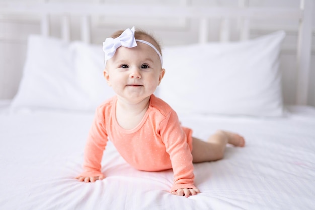 Una niña caucásica feliz con un traje rosa yace en la cama de su casa en el dormitorio con ropa de cama blanca mira a la cámara y se ríe de un bebé saludable