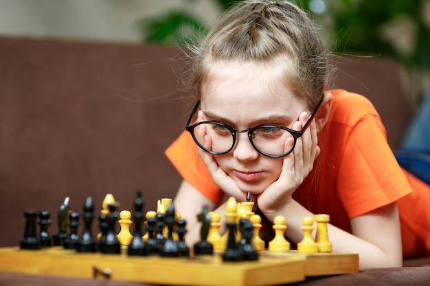 Foto niña caucásica con gafas pensando en un juego de ajedrez jugando al ajedrez en casa durante la cuarentena. el desarrollo de la primera infancia.